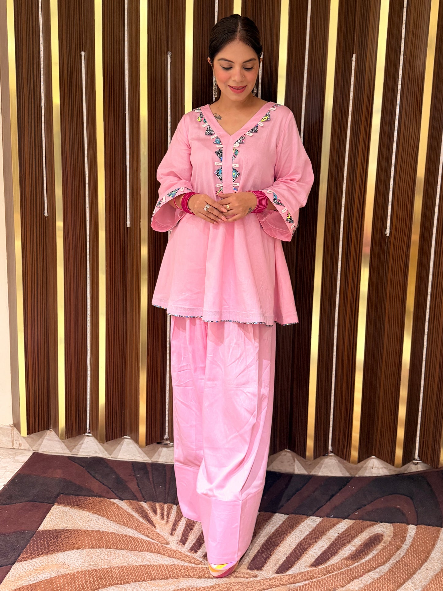 A woman wearing a pink traditional outfit with embroidered details on the neckline and sleeves, standing in front of a vertical wood slat wall.